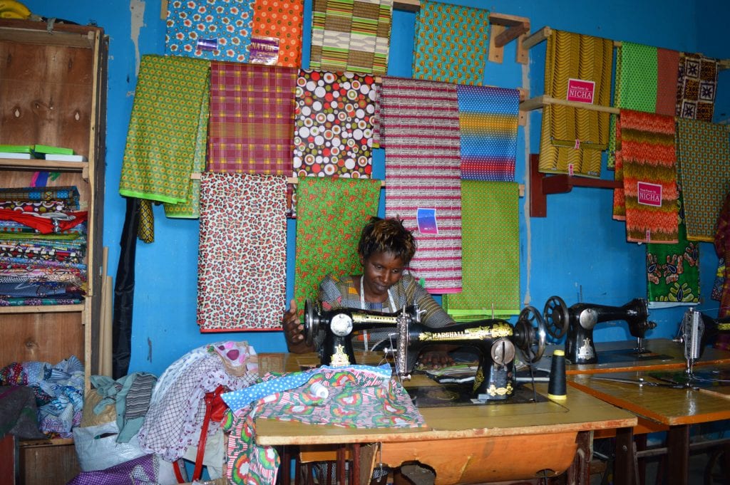 Young woman operating a sewing machine in a colorful workshop, surrounded by vibrant fabrics hanging on the walls and shelves stocked with textiles.