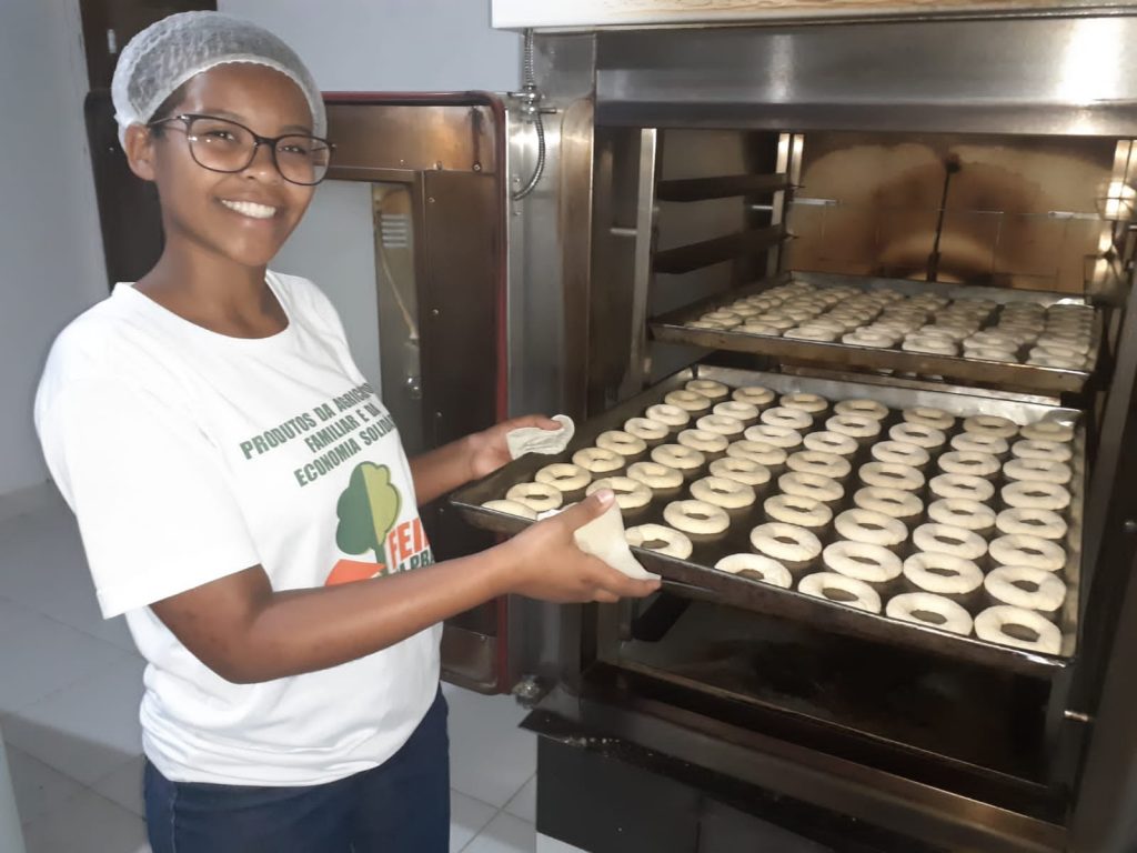 Smiling young woman wearing a hair net and glasses, placing a tray of small, round baked goods into a large industrial oven.