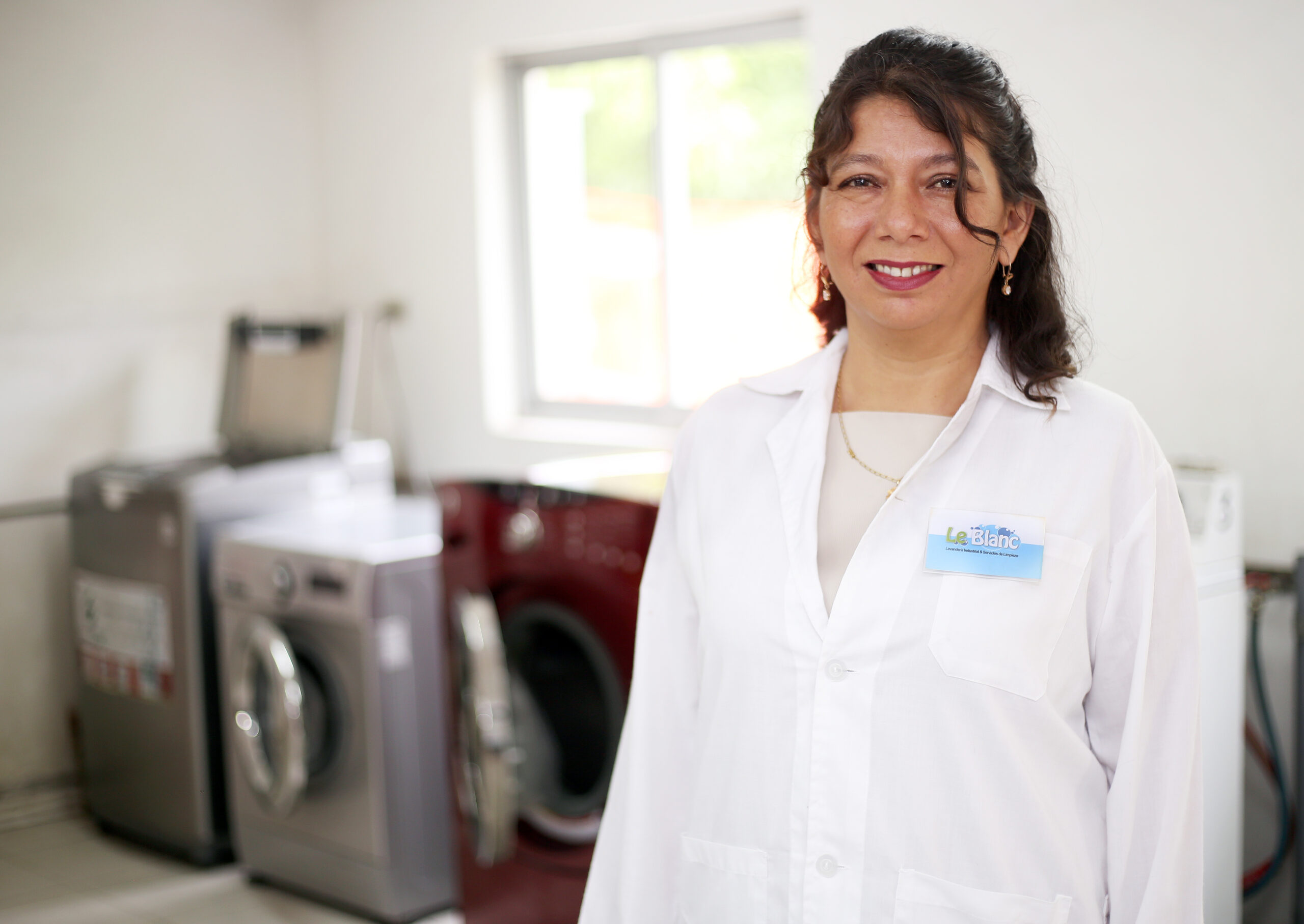 A woman stands on the right, wearing a white jacket, and smiling at the camera. There are several washing machines in the background. She is Jessica Vargas, an entrepreneur who successfully expanded her laundry business thanks to training from TechnoServe. 