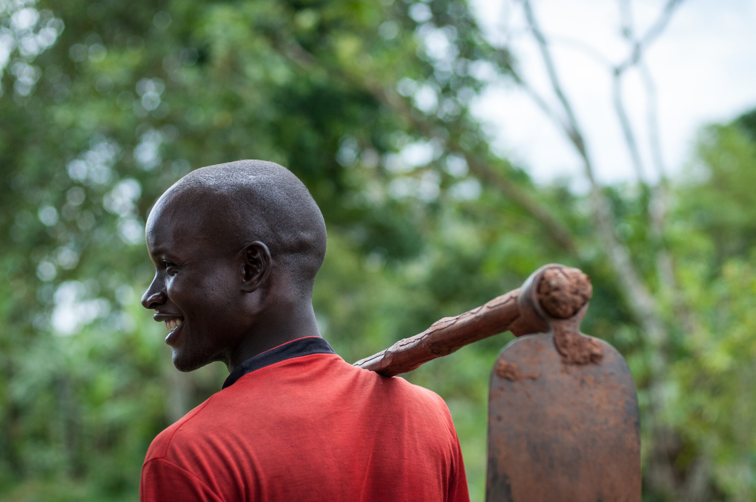 A young man is seen smiling in profile against a lush green background. He carries a muddy hoe on his shoulder. This is Yossam Mukobba, a farmer in Uganda, who received training from TechnoServe. 