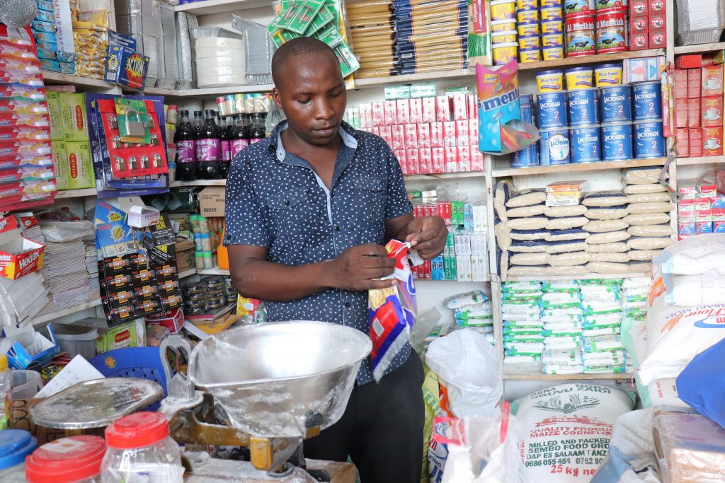 Young man in a blue patterned shirt standing behind the counter of a small, well-stocked convenience store filled with various household goods and food items. Part of a feature for World Youth Skills Day. 