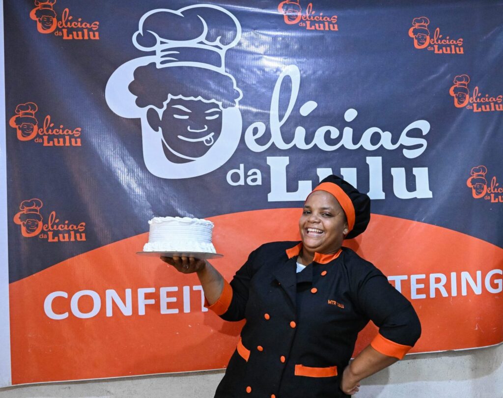 A woman holds a cake in one hand and smiles at the camera against a backdrop that reads Delicias da Lulu. She is Lúcia Sigauque, an entrepreneur in Mozambique who has made a success of her bakery with training support from TechnoServe. 