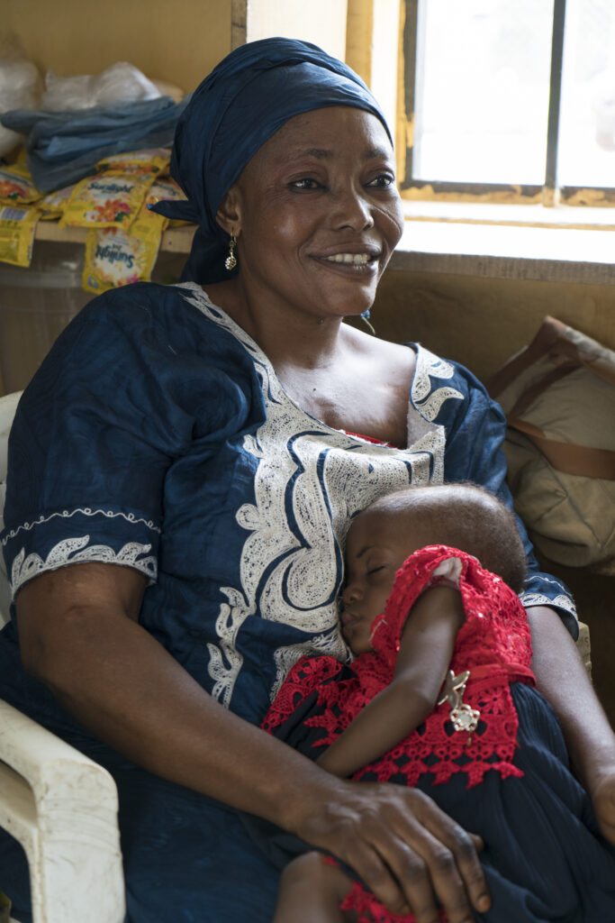 A healthy young baby sleeps in their mother's arms. She is smiling and sitting on a chair. About 11 million young children in Nigeria are struggling with severe food poverty, making them 50% more likely to suffer from wasting according to UNICEF.  