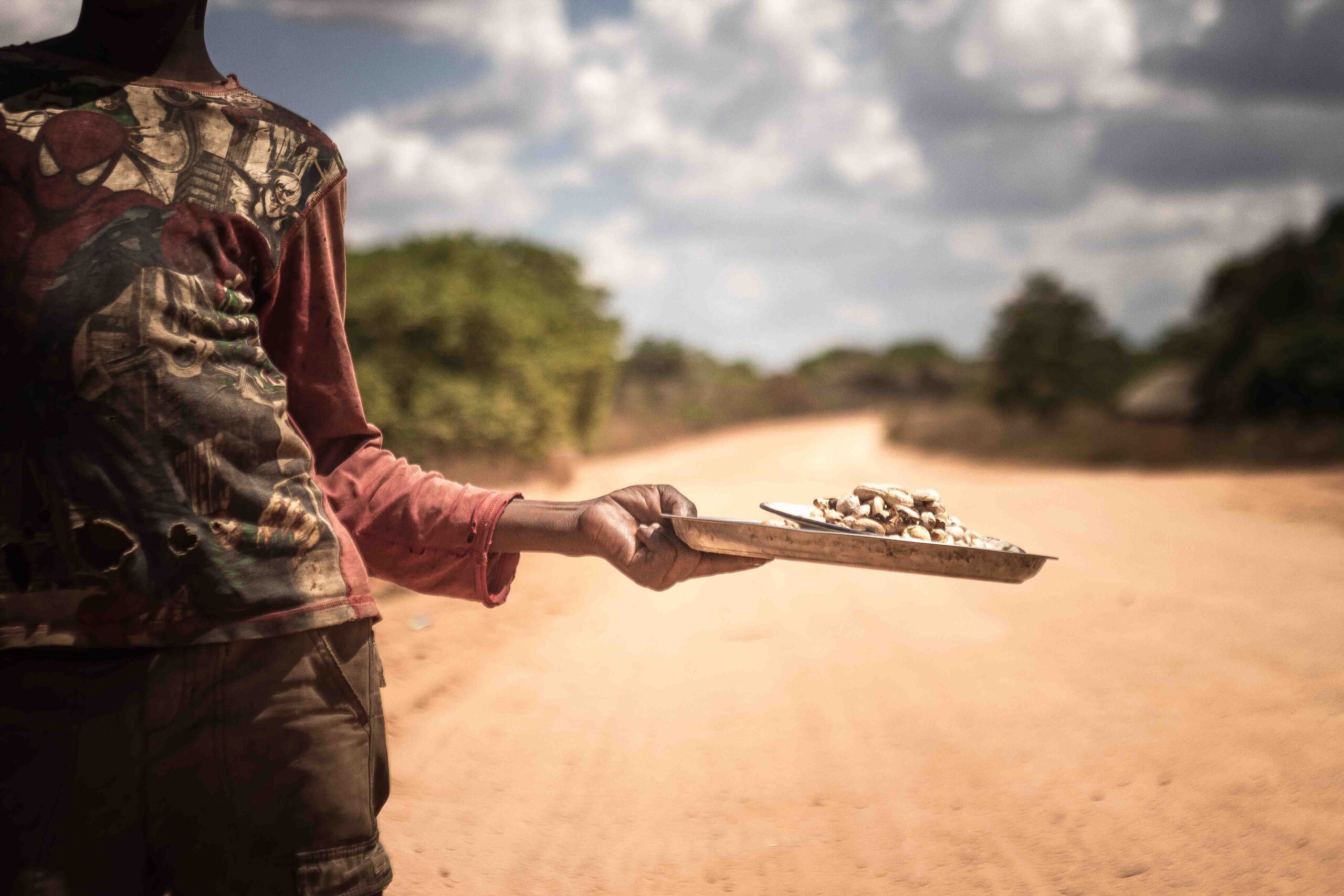 A person holding a tray of cashew extended in one hand, standing on a dirt road under a cloudy sky.